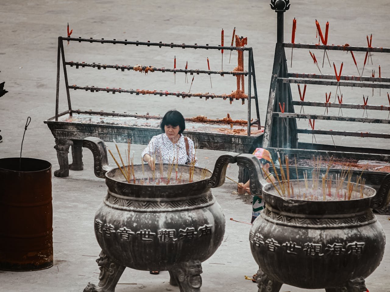 An adult Asian woman praying at a temple, surrounded by large incense burners emitting smoke.