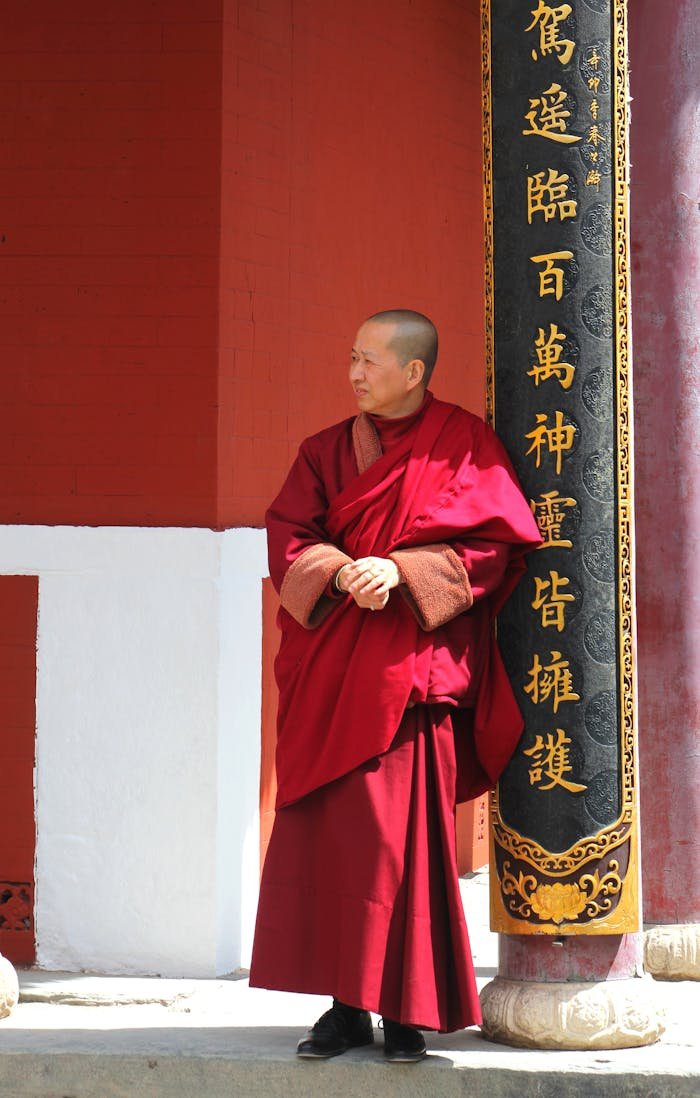 A Buddhist monk in traditional attire stands at a temple with Chinese inscriptions.